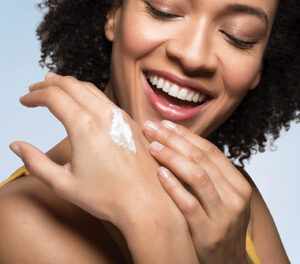 Image of woman smiling with her hands covering her chin slightly. Close up on hands shows a cream swash on top of one hand indicating the other hand is spreading it.