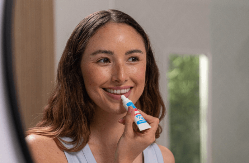 Promotional image showing a woman smiling while applying Flexitol Lip Balm Original to her lips
